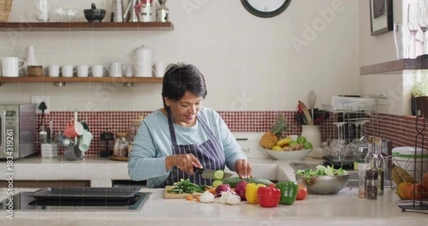 Fototapeta Chopping senior Indian cook in striped apron slicing cucumber at kitchen island, with cutting board