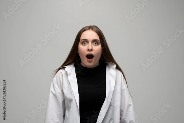 Fototapeta Young woman looking shocked with wide eyes and open mouth in studio portrait on gray background