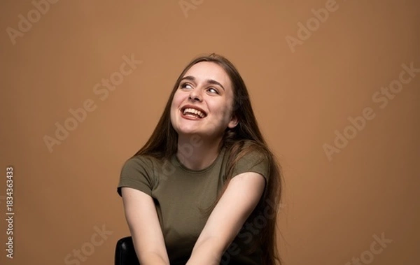 Fototapeta Smiling young woman looking up with joyful expression on brown background, positive human emotion portrait