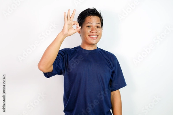 Fototapeta A cheerful young Asian man in a blue shirt stands against a clean white background, smiling and giving an OK gesture.