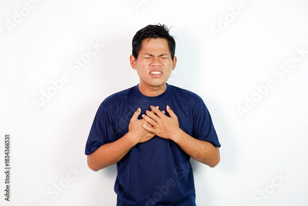 Fototapeta A young Asian man in a blue shirt clutches his chest with a pained expression, conveying distress or chest discomfort in a simple studio setting.