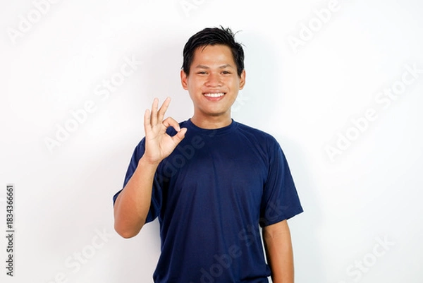 Fototapeta A cheerful young Asian man in a blue shirt stands against a clean white background, smiling and giving an OK gesture.