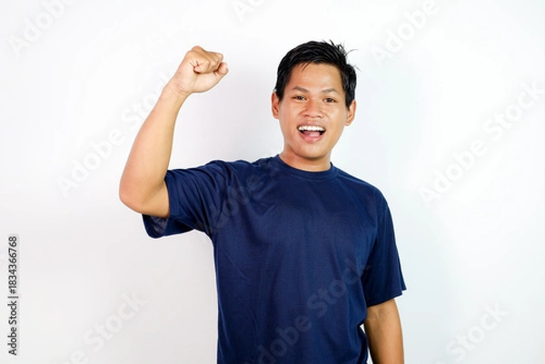 Fototapeta Happy Young Man In Blue T‑Shirt Celebrating Victory With Raised Fists and A Wide Open Mouth, Energetic Expression.