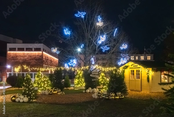 Obraz A festive Christmas park scene with glowing trees, illuminated angel decorations, ornaments, and a warmly lit holiday cabin at night.