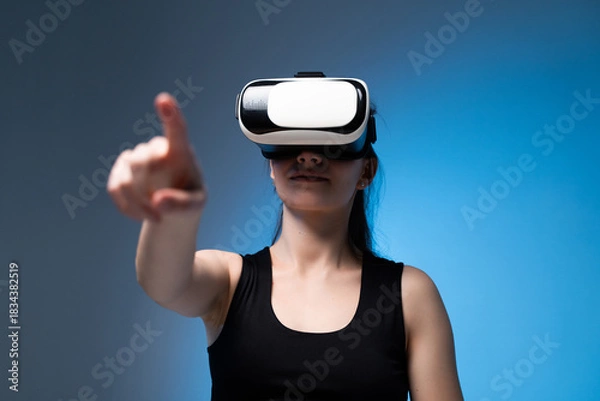 Fototapeta Young woman reaching forward while using VR headset in immersive digital environment with blue lighting