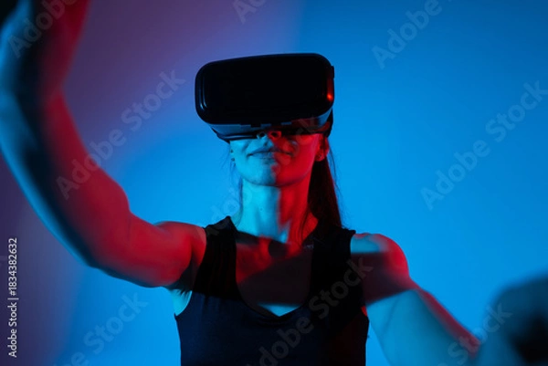 Fototapeta Woman raising arms while wearing VR headset and interacting with virtual elements in neon environment