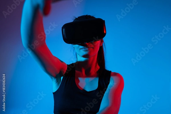 Fototapeta Woman engaged in virtual reality experience wearing VR headset in vivid blue red neon futuristic lighting
