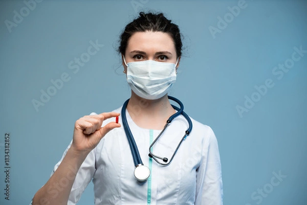 Fototapeta Female doctor in medical mask showing red pill toward camera while wearing white uniform