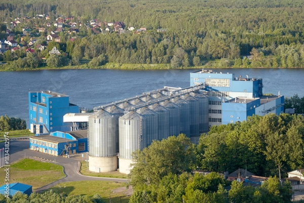 Obraz A factory stands near a river with large silos, surrounded by trees and houses. The scene shows clear skies and bright sunlight during the day. People may work inside the factory
