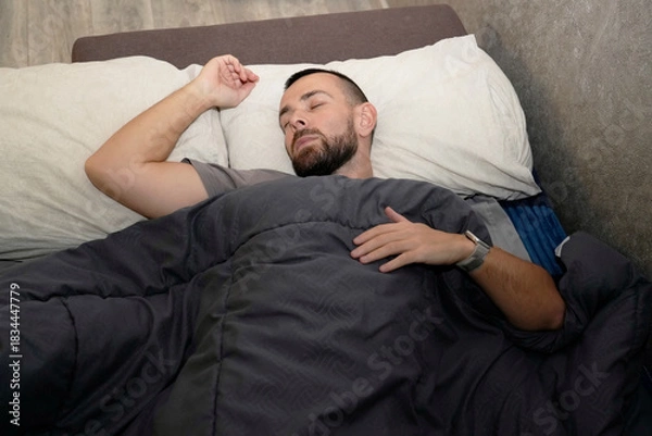 Obraz A man lies in bed under a blanket, appearing to sleep. His face shows signs of fatigue. It seems he is taking time off for recovery at home during the daytime