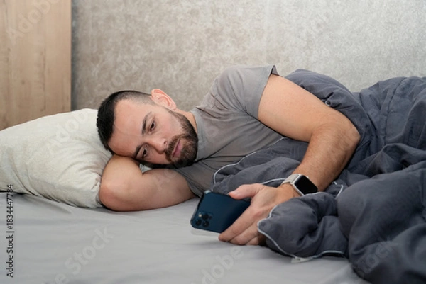 Obraz A man lies in bed under a blanket, checking his phone. He looks tired and is on sick leave to recover from an illness. The time is likely during the day