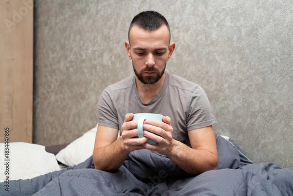 Obraz A man sits in bed holding a mug, looking tired and unwell. He is resting under a blanket, taking time off work to recover from an illness and regain his health