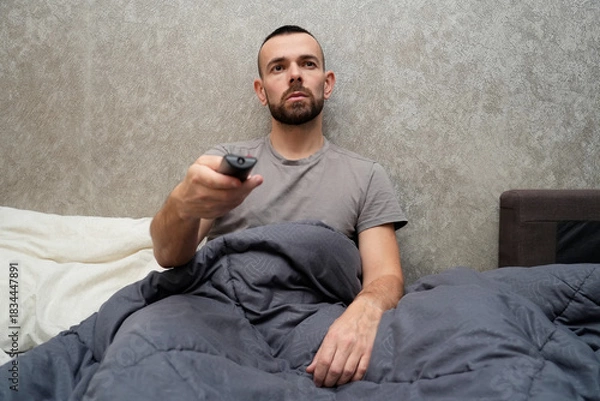 Obraz A man sits up in bed, wrapped in a blanket, holding a TV remote. He looks tired and is taking time off work to rest and recover from illness at home