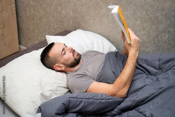 Obraz A man lies in bed with a blanket, reading a book. He shows signs of fatigue as he takes time to recover from an illness while resting at home