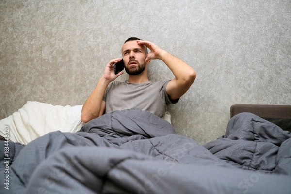 Obraz A man lies in bed covered with a blanket, appearing fatigued. He talks on the phone, seeking help or sharing updates while resting in his bedroom at home during the day