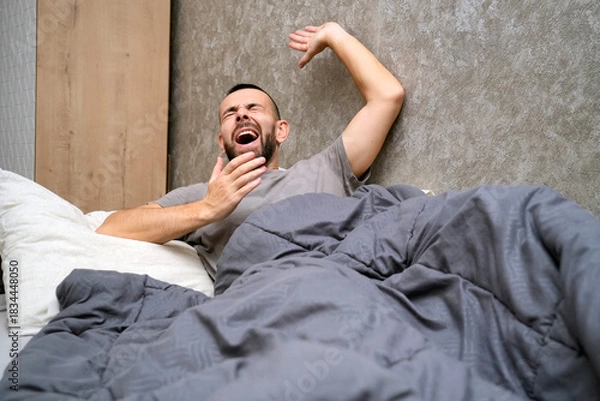 Obraz A man lies in bed under a blanket, yawning widely. He appears to be experiencing fatigue and is taking time to recover from an illness at home in a simple room