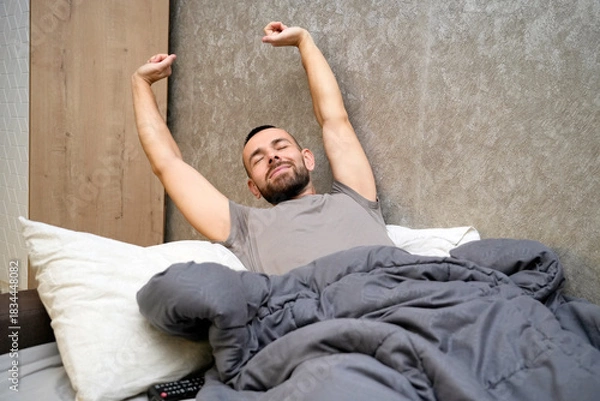 Obraz A man is waking up in bed, stretching his arms above his head. He has a relaxed expression on his face. The blanket is draped over him as he enjoys a moment of rest after being unwell