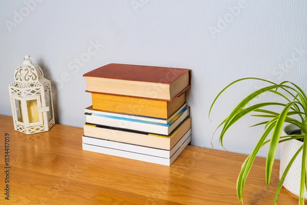 Fototapeta Pile of books on a cabinet neatly stacked on top of each other with a lantern candle holder and a spider plant, reading or library based activity concept