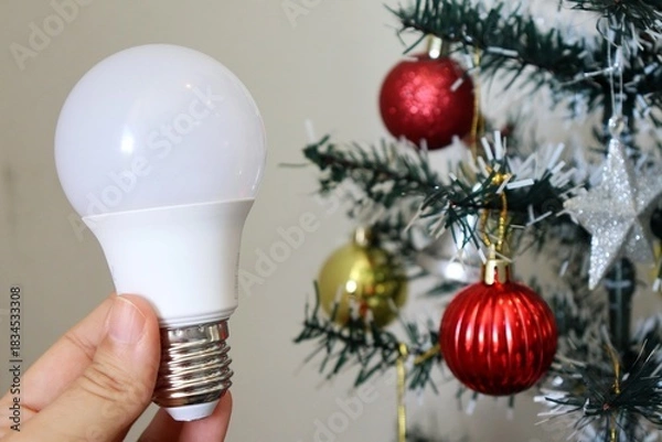 Obraz Hand holding LED light bulb with decorated Christmas tree in background. Electricity bill concept during the Christmas holiday. 
