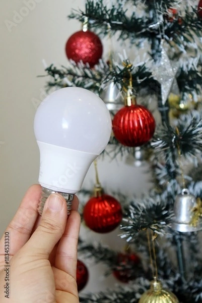 Obraz Hand holding LED light bulb with decorated Christmas tree in background. electricity bill concept during the Christmas holiday. 