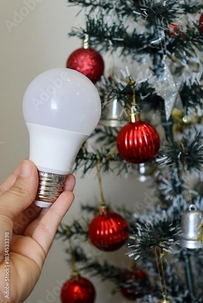 Obraz Hand holding LED light bulb with decorated Christmas tree in background. Electricity bill concept during the Christmas holiday. 