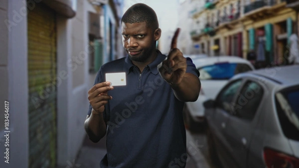 Fototapeta Young black man holds creditcard and points finger at camera in narrow street with parked cars and shuttered storefronts; warning security trust.