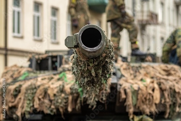 Fototapeta Close-up of a camouflaged tank cannon pointing towards the sky in an urban setting, with building with soldiers walking on the turret