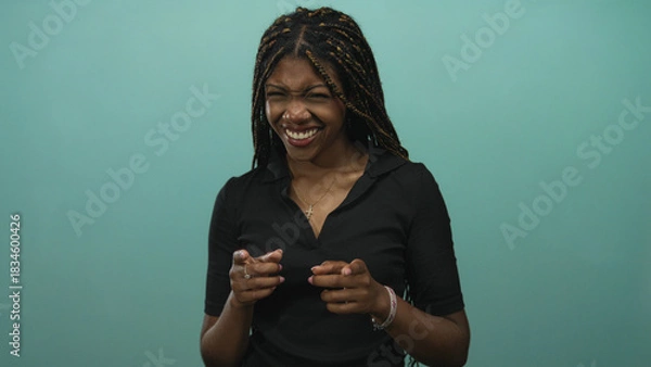 Fototapeta Woman pointing finger and smiling in teal studio, braided hair and hands gesturing toward camera; confidence playfulness warmth.