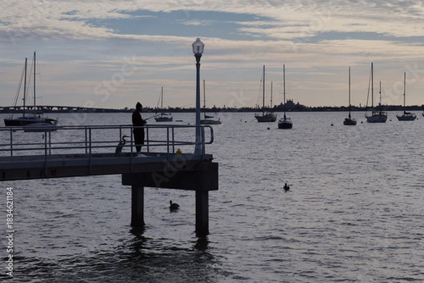 Obraz Harbor Pier At Dusk With Silhouette Fisherman, Sailboats And Calm Water Scene in Gulfport, FL. Distant sailboats. Gentle mood with subtle light and reflective surface.
