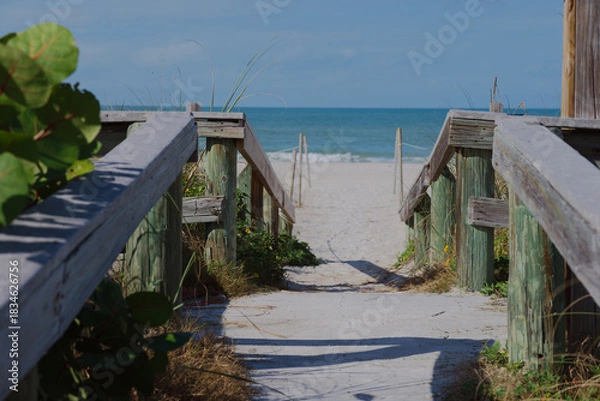 Obraz Sunny Beach Path With Weathered Wooden Railings Leading To Quiet Sea View at St. Pete Beach, FL. Sandy path opening to a calm blue sea. Ideal for travel, coastal lifestyle, relaxation, and nature them