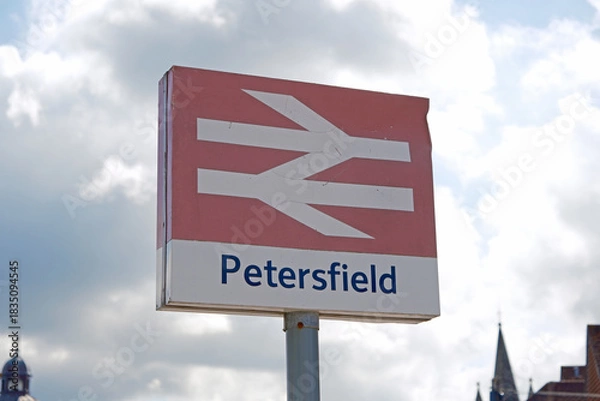 Fototapeta Petersfield Station Sign with National Rail Logo Against Cloudy Sky and Distant Buildings