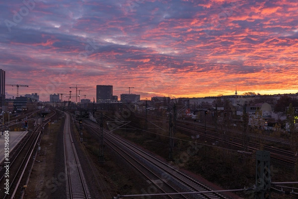 Obraz Railway tracks curve into the distance under a vivid red-orange dawn sky. Urban skyline and construction cranes frame a moody Berlin morning.