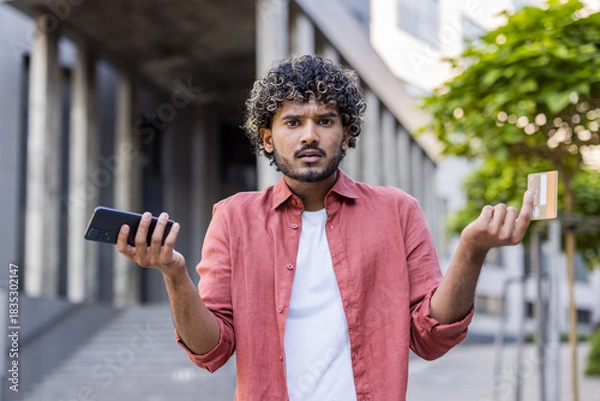 Fototapeta Close-up portrait of a young worried Indian man standing outside having problems with his credit card and phone