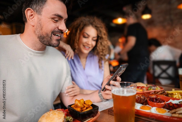 Fototapeta Couple laughing and using smartphone while having lunch at restaurant