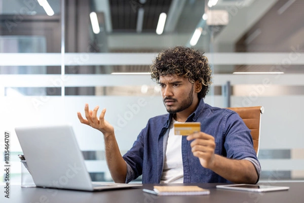 Fototapeta Frustrated young Indian man found an error and problem with his bill and credit card which he holds in his hand and looks at his laptop