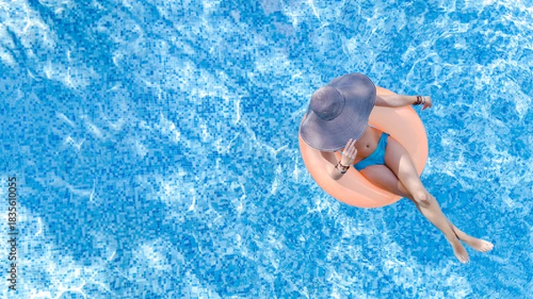 Fototapeta Aerial drone view of beautiful woman in hat relaxing in swimming pool, young girl in bikini swims on inflatable ring donut and has fun in blue pool water, tropical vacation on holiday resort