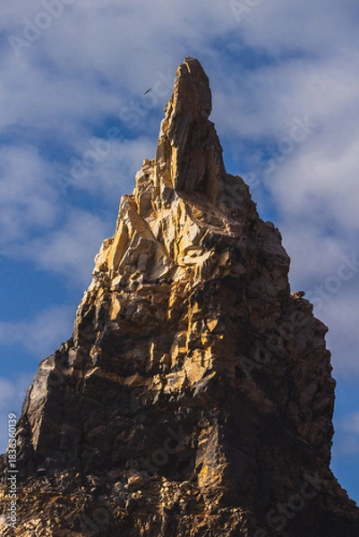 Fototapeta Sharp Rocky Peak with Bird in Flight 
A dramatic rocky pinnacle illuminated by warm sunlight, with a solitary bird gliding above against a soft, cloudy sky.