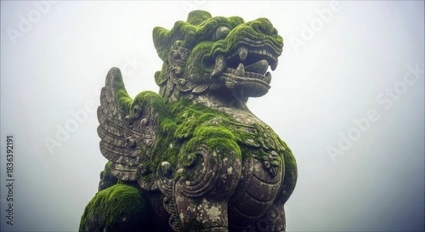 Fototapeta A weathered stone dragon statue covered in green moss, set against a backdrop of a foggy sky.