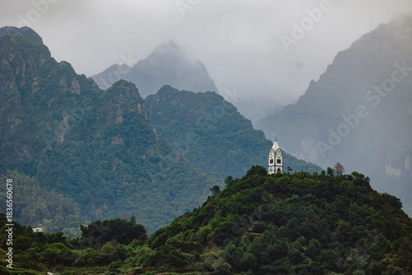 Obraz Scenic view of a small white church perched on a hill surrounded by lush green mountains and misty clouds, creating a serene and tranquil atmosphere in nature