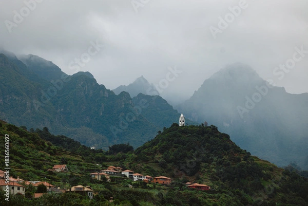 Obraz Lush green hills with scattered colorful houses beneath dramatic mountains shrouded in mist, showcasing a serene landscape and natural beauty in a tranquil environment