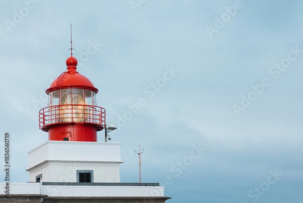 Obraz Red and white lighthouse with a glass lantern, standing tall against a cloudy sky, symbolizing guidance and safety for maritime navigation and coastal scenery