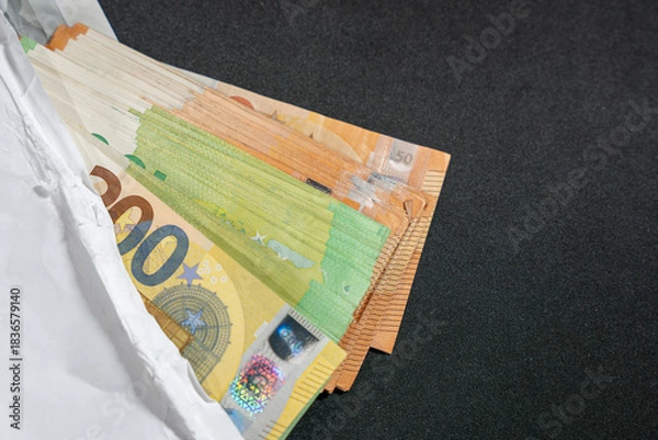 Fototapeta A white envelope reveals a stack of euro banknotes on a dark background. Symbol of salary, gift money, saving, or hidden cash.