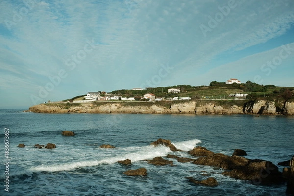 Fototapeta Northern Spain coastal landscape. Ocean with small waves and rocks in the foreground, a high coastal cliff with a walking trail and low hills with scattered houses in the background near Gijon