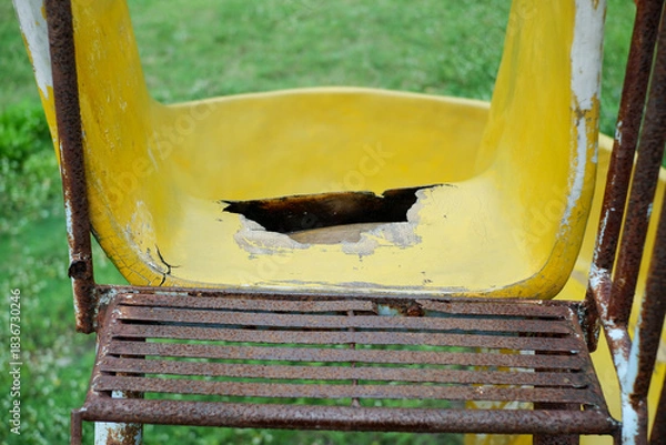 Fototapeta Close-up of a weathered yellow playground slide showing chipped paint, a large hole in the seat area, and a rusted metal frame. Great for themes of decay, neglect, and worn outdoor playgrounds.