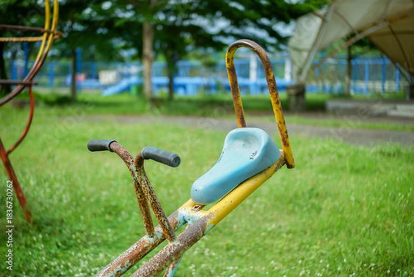 Fototapeta Rusty seesaw with blue seat in green park playground. A faded, rusted seesaw stands in a grassy park, its blue seat and yellow frame telling a story of long use.