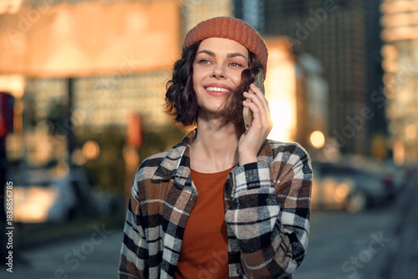 Obraz woman on phone with smile in urban street, candid lifestyle portrait at golden hour glow; beanie and plaid jacket convey authenticity, mindful living and emotional storytelling, soft warm light and