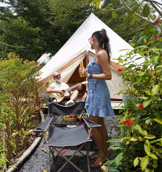 Obraz A man and woman is joyfully drinking beer and playing guitar near a cozy tent at glamping