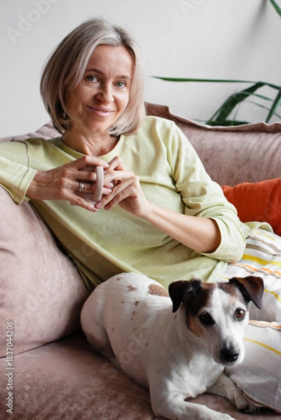 Obraz Happy middle aged Woman sitting with Dog on Sofa in living room with cup of tea or coffee