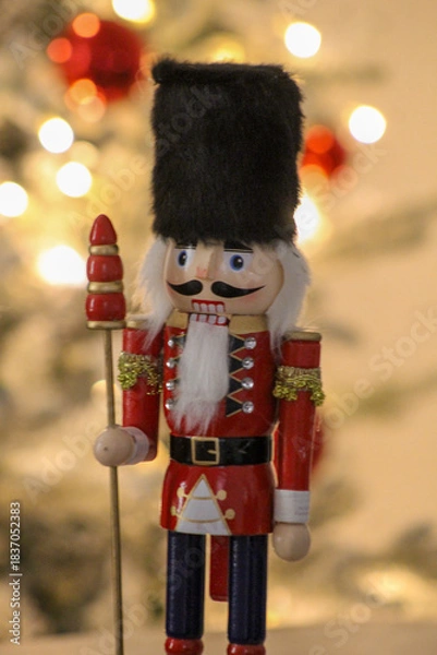 Obraz Vertical image of a colorful nutcracker figure placed near a glowing Christmas tree with red ornaments.