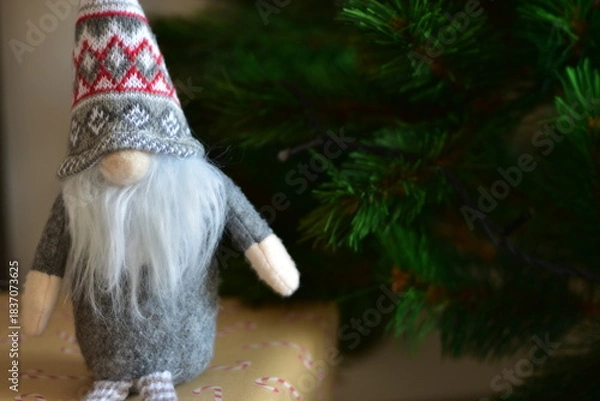 Fototapeta Cozy Christmas scene with a small Nordic gnome sitting on wrapped gifts under a decorated tree. Kraft paper boxes, soft handmade toy and natural holiday decor create a warm festive atmosphere at home.